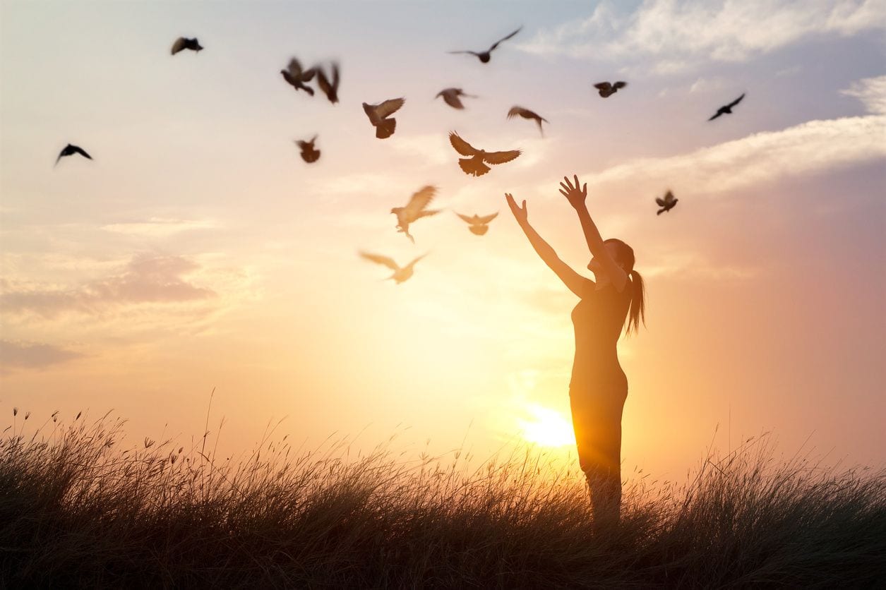 Woman with her arms raised as sun sets and birds flying