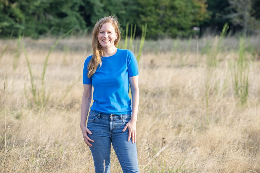 Jennifer smiling and wearing a blue shirt and jeans while standing in a grassy field