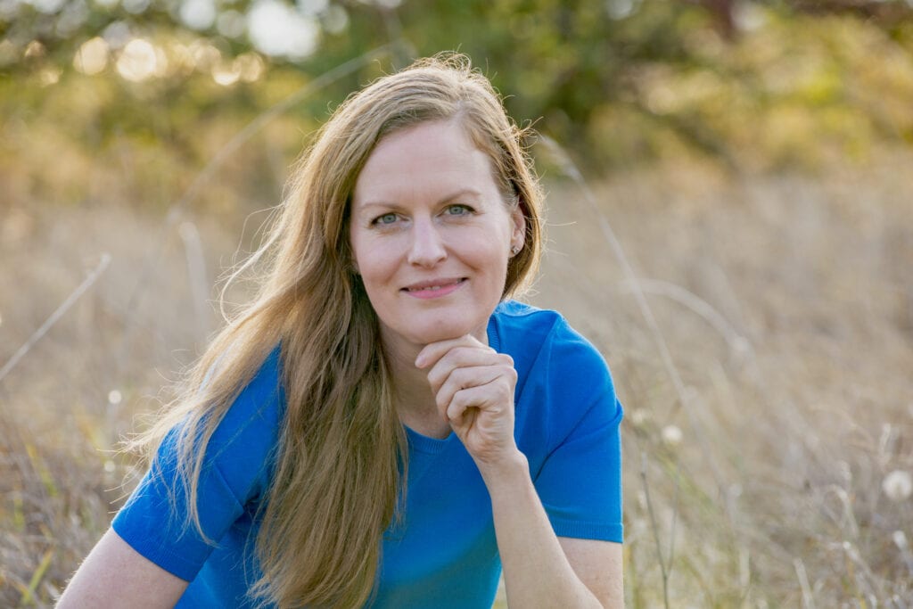 Jennifer smiling and wearing a blue shirt and jeans while sitting in a grassy field